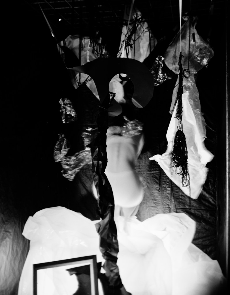 The image depicts a woman in a black dress and hat, surrounded by various objects, including a picture frame, in a dark room.